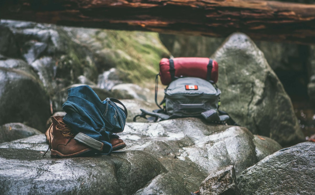 Outdoor Camping Szene mit robustem Motorrad-Campingzubeh&ouml;r: Wanderschuhe, gefaltete Jeans und ein Rucksack mit Isomatte auf Felsen in nat&uuml;rlicher Umgebung.