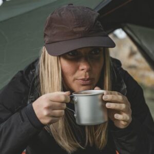 Frau mit brauner Kappe hält eine silberne Aluminiumausstecher-Tasse während Motorrad-Camping.