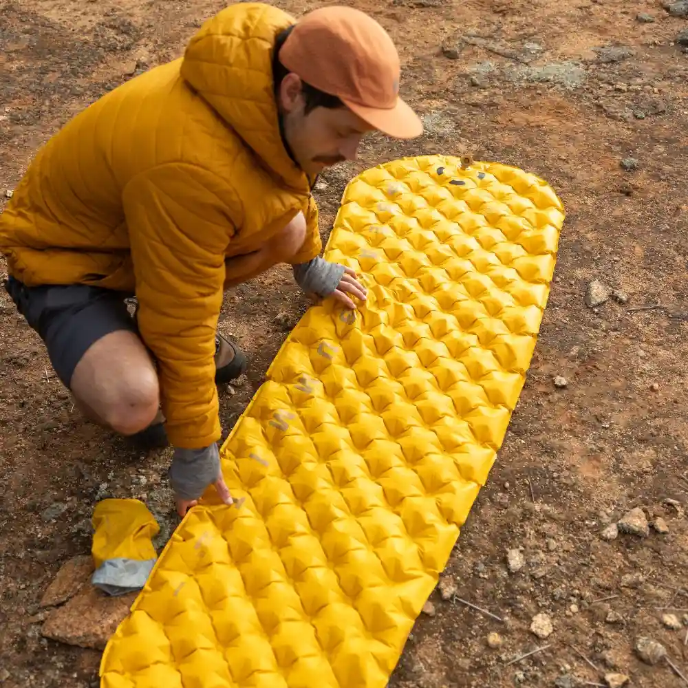 Mann in gelber Jacke und orangener Mütze richtet eine gelbe, ultraleichte XR Isomatte für Motorrad-Camping aus. Die Isomatte befindet sich auf erdigem Boden, ideal für Outdoor- und Camping-Aktivitäten.