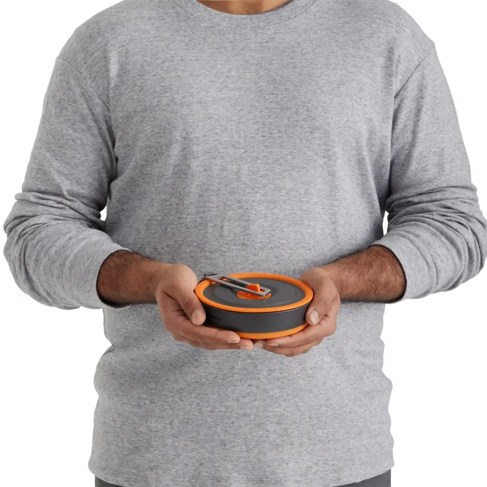 Ein Mann in grauem Pullover hält ein kompaktes, rundes 3-teiliges Kochset mit orangen Akzenten in den Händen, ideal für Motorrad-Camping und Outdoor-Aktivitäten.
