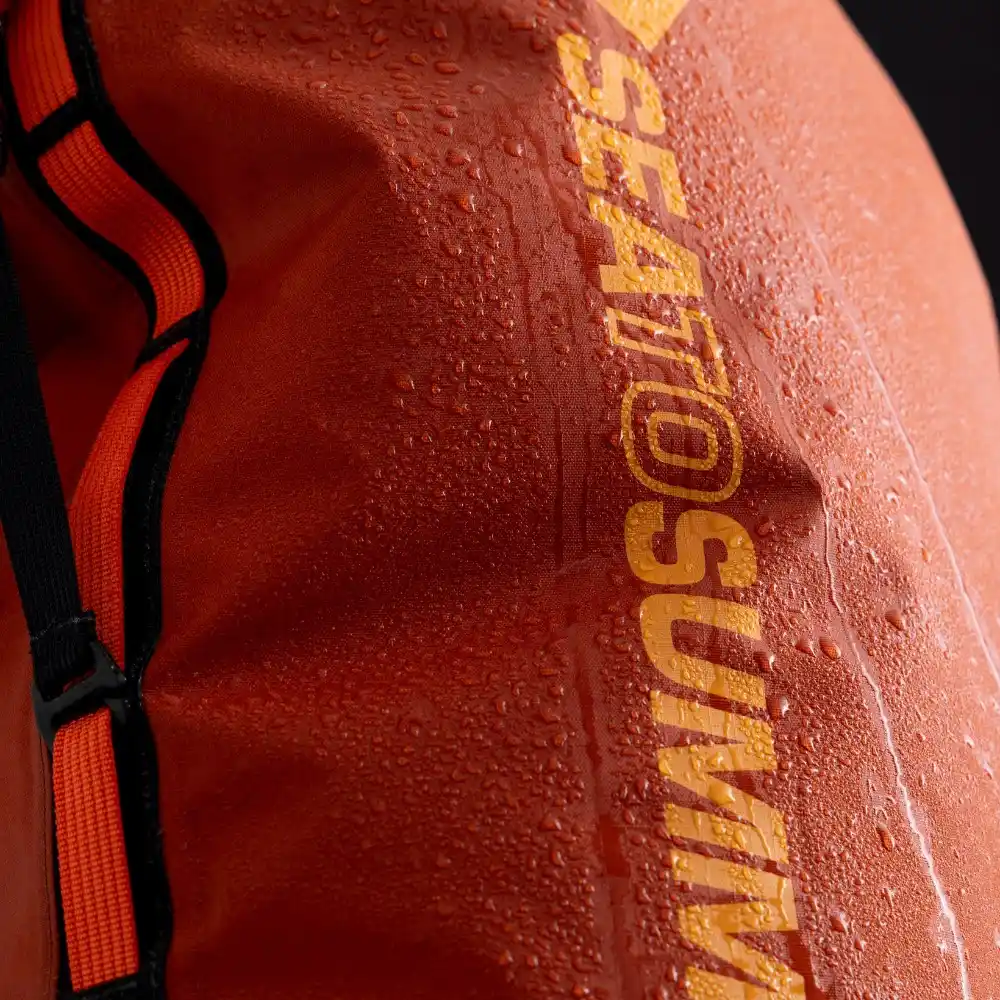 Nahaufnahme eines orangefarbenen Big River Dry Backpacks von Sea to Summit, der mit Wassertropfen bedeckt ist, betont die wasserdichte Beschaffenheit des 50-Liter-Rucksacks, ideal für Motorrad-Camping-Abenteuer.
