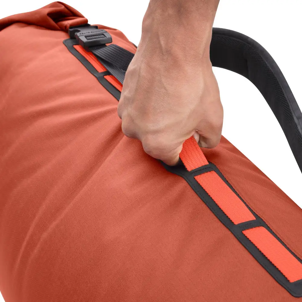 Nahaufnahme eines Big River Dry Backpack 50 Liter in Rostfarbe, der von einer Hand am Griff gehalten wird. Ideal für Motorrad-Camping, bietet der wasserdichte Rucksack robusten Schutz für Outdoor-Ausrüstung.
