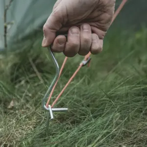 Nahaufnahme einer Hand, die einen Zelthering in der Erde befestigt, mit robustem Seil - essenzielles Zubehör für Motorrad Camping 2026.