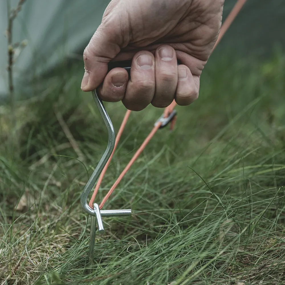 Nahaufnahme einer Hand, die einen Zelthering in der Erde befestigt, mit robustem Seil - essenzielles Zubehör für Motorrad Camping 2026.