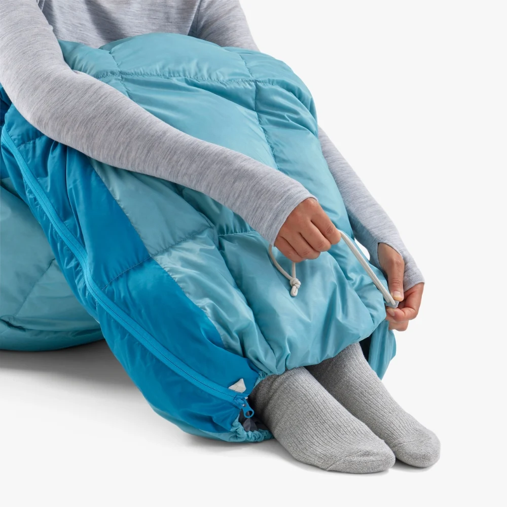 Person in einem hellblauen Daunenschlafsack mit grauer Kleidung und Socken beim Motorrad-Camping. Der Schlafsack hat eine Kordelzugöffnung, die von der Person angepasst wird.