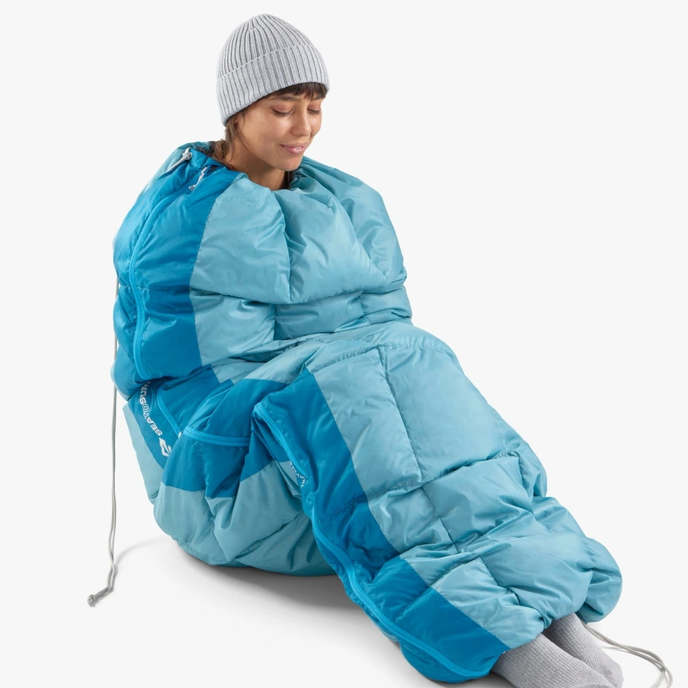 Frau sitzt in einem blauen Daunenschlafsack und trägt eine graue Strickmütze, ideal für Motorrad-Camping.